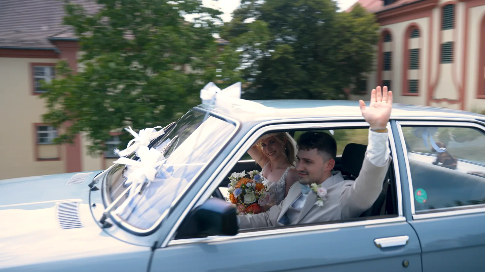 Das Brautpaar winkt aus dem Oldtimer vor dem Schloss